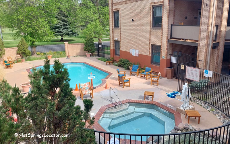 Outdoor pool area of Best Western Plus Plaza Hotel in Thermopolis, Central Wyoming. The area features a swimming pool with clear blue water, a smaller hot tub, and wooden lounge chairs with umbrellas. The setting is illuminated by warm outdoor lighting, providing a relaxing ambiance.