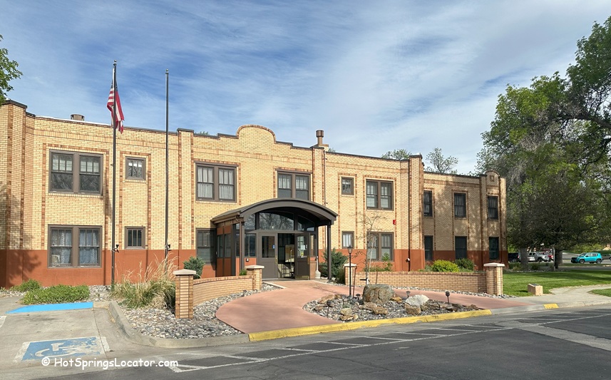 Best Western Plus Plaza Hotel in Thermopolis, Central Wyoming. The hotel is a two-story brick building with large windows, surrounded by lush green trees and grass. A prominent sign displaying the Best Western Plus Plaza Hotel logo is visible in front of the building.