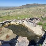 Prince Albert Hot Spring - Idaho: A natural hot spring pool lined with rocks, filled with clear water, and set in a grassy, open landscape with rugged hills and blue sky in the distance.
