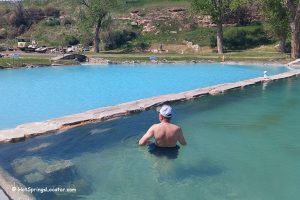 Fountain of Youth RV Park in Thermopolis Wyoming: A man sits and relaxes in a hot spring pool with clear greenish water, separated by a stone wall from a larger blue pool, surrounded by grassy areas and trees.