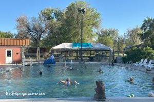 Hellie's TePee Pools and Spa at Hot Springs State Park in Thermopolis Wyoming: Outdoor swimming pool with people relaxing in the water, white lounge chairs on the deck, and a white and blue canopy tent with trees behind.