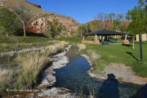 Hot Springs State Park in Thermopolis Wyoming: A clear hot spring stream winds through grassy parkland, with a picnic shelter and red rocky hills in the distance.