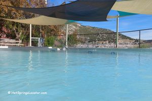 State Bath House at Hot Springs Park in Thermopolis Wyoming: Outdoor hot springs pool with light blue water, shade sails overhead, and mountain views beyond the fence.
