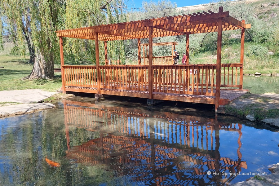 Indian Springs Resort and RV in Idaho: A wooden pergola bridge stretches over a clear pond, with reflections in the water and willow trees and grassy lawns in the background.