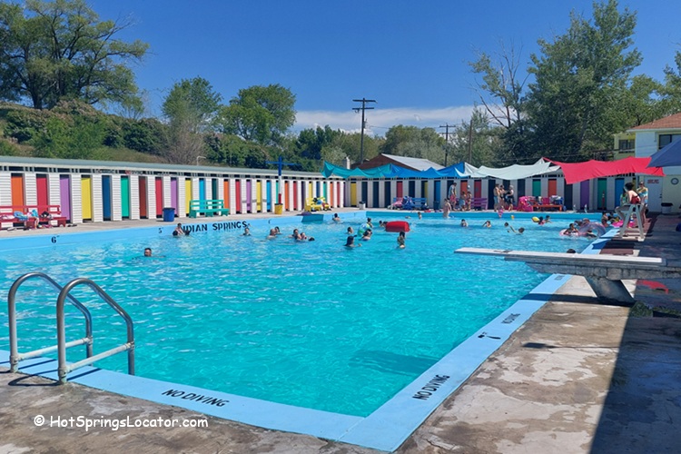 Indian Springs Resort and RV in Idaho: An outdoor pool with swimmers and pool toys, surrounded by colorful cabanas and shade canopies, with trees and clear blue sky in the background.