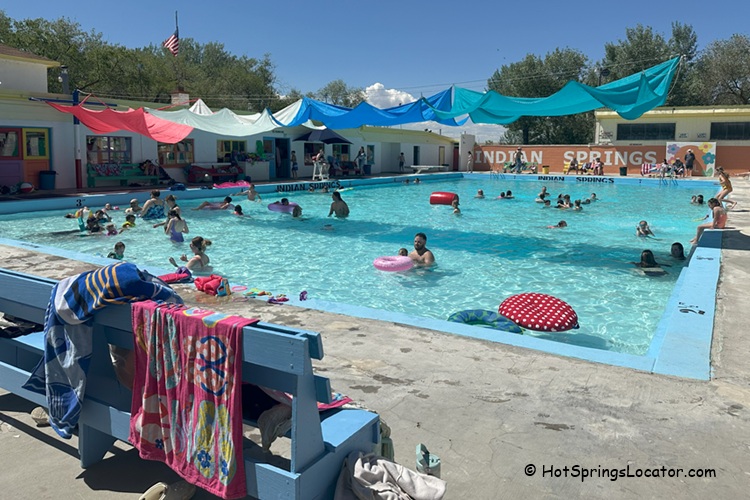Indian Springs Resort and RV in Idaho: A large outdoor swimming pool filled with people and colorful floaties, with towels hanging over a blue bench and shade sails overhead.