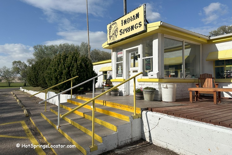 Indian Springs Resort and RV in Idaho: The entrance to the resort features a white building with yellow trim, a sign reading "Indian Springs," yellow handrails, and outdoor seating surrounded by trees.
