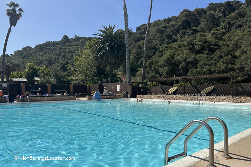Avila Hot Springs in California: The picture "Avila Hot Springs in California swimming pool" shows a spacious outdoor swimming pool under a clear blue sky, with a few swimmers in the water, palm trees and hills in the background, and lounge chairs along the fence.