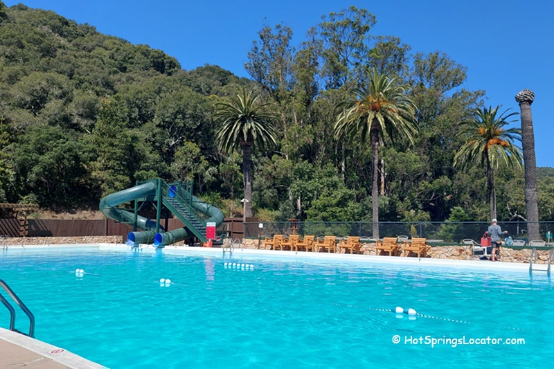 Avila Hot Springs in California: The picture "Avila Hot Springs in California swimming pool and slide" features a large outdoor swimming pool with clear blue water, a green water slide in the distance, palm trees, lounge chairs, and a forested hillside behind the pool.