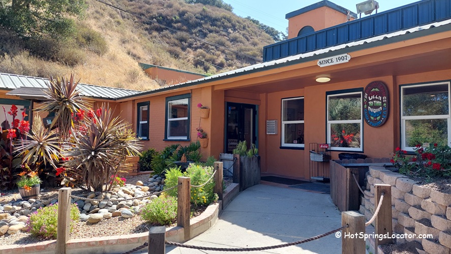 Avila Hot Springs in California: The picture "Avila Hot Springs in California entrance" shows the front entrance to Avila Hot Springs, with an orange stucco building, a welcoming sign, desert landscaping with rocks and spiky plants, and a hillside in the background.