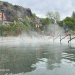 Lava Hot Springs Foundation World Famous Hot Springs Idaho: The picture "Lava Hot Springs Foundation World Famous Hot Springs Idaho steaming pool by cliffs" shows a steaming hot spring pool with handrails, set against rocky cliffs and greenery, with American flags and colorful banners on lamp posts.