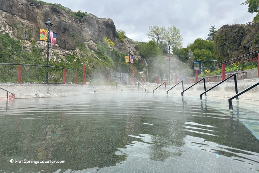 Lava Hot Springs Foundation World Famous Hot Springs Idaho: The picture "Lava Hot Springs Foundation World Famous Hot Springs Idaho steaming pool by cliffs" shows a steaming hot spring pool with handrails, set against rocky cliffs and greenery, with American flags and colorful banners on lamp posts.