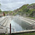 Lava Hot Springs Foundation World Famous Hot Springs Idaho: The picture "Lava Hot Springs Foundation World Famous Hot Springs Idaho main pool with stone steps" presents a large rectangular hot spring pool with steam rising, flanked by wide stone steps and retaining walls, with rocky hills and greenery in the background.