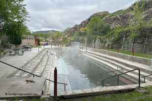 Lava Hot Springs Foundation World Famous Hot Springs Idaho: The picture "Lava Hot Springs Foundation World Famous Hot Springs Idaho main pool with stone steps" presents a large rectangular hot spring pool with steam rising, flanked by wide stone steps and retaining walls, with rocky hills and greenery in the background.