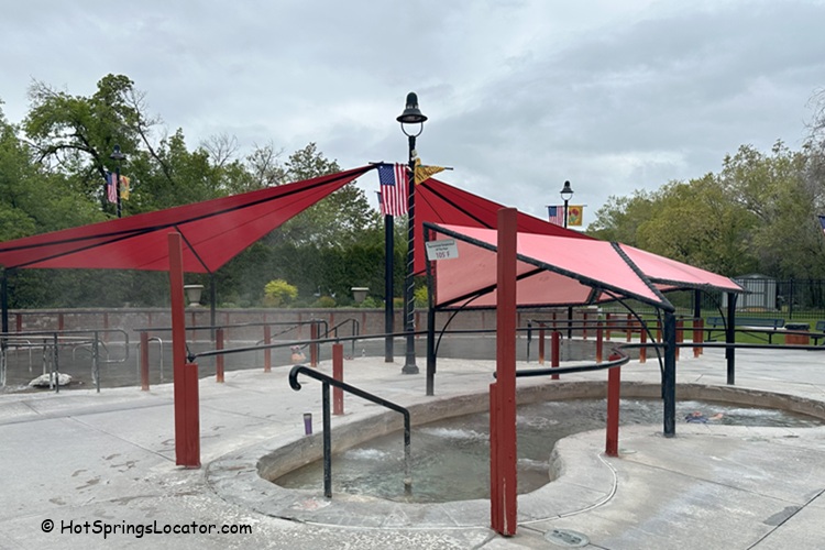 Lava Hot Springs Foundation World Famous Hot Springs Idaho: The picture "Lava Hot Springs Foundation World Famous Hot Springs Idaho small covered soaking pool" shows a small circular soaking pool with railings, shaded by red canopies, and surrounded by paved paths, lamp posts, and green lawns.
