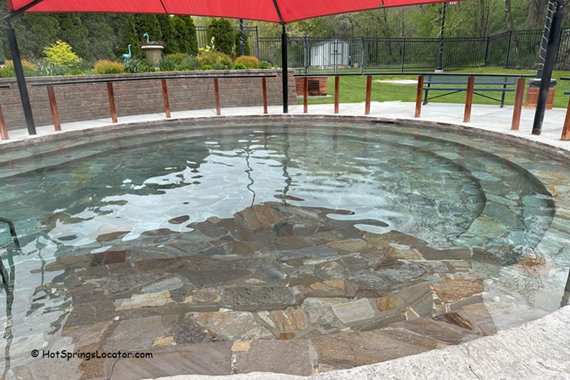 Lava Hot Springs Foundation World Famous Hot Springs Idaho: The picture "Lava Hot Springs Foundation World Famous Hot Springs Idaho round stone pool" displays a circular stone hot spring pool with clear water, covered by a red canopy, surrounded by a landscaped garden and open picnic area.