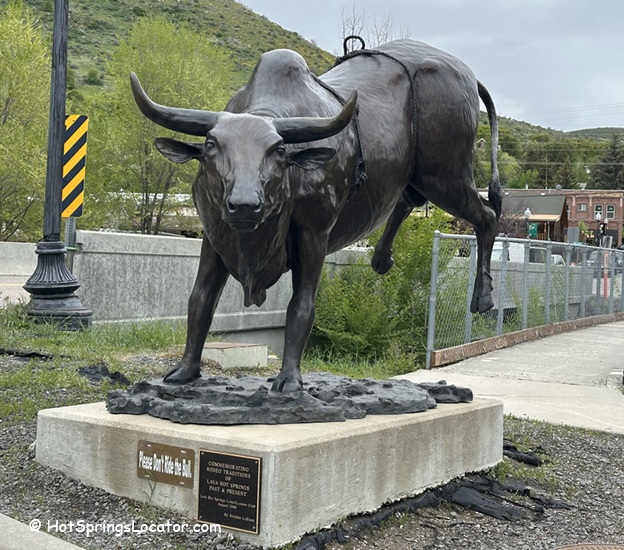 The Town of Lava Hot Springs Idaho: The picture "The Town of Lava Hot Springs Idaho bull statue" features a large bronze bull statue on a concrete base, with a plaque in front, set beside a sidewalk with green hills and buildings in the background.