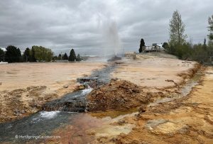 Soda Springs Geyser in Idaho - A close-up of the geyser eruption with water shooting high into the air against a cloudy sky. The foreground features mineral-rich ground, and a small structure is visible to the right in the background.