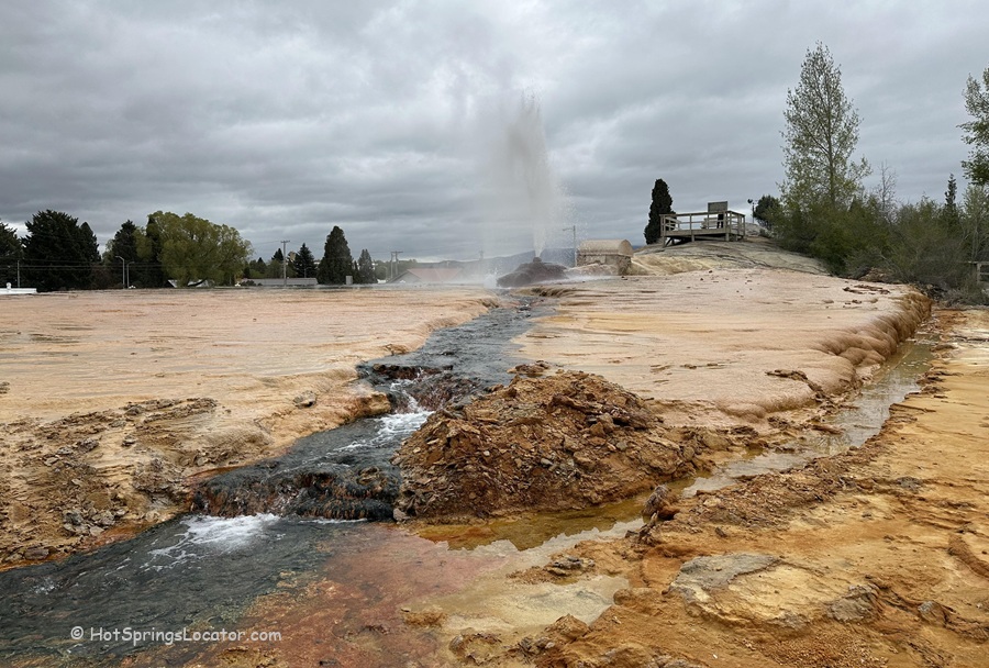 Soda Springs Geyser in Idaho - A wide view of the geyser erupting, with a stream of mineral-rich water flowing through orange and yellow ground. A wooden viewing platform and trees are visible in the distance under a cloudy sky.