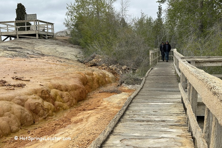 Soda Springs Geyser in Idaho - A wooden boardwalk leading through mineral deposits and trees, with two people walking in the distance. To the left, orange and yellow mineral formations are visible, blending into the natural surroundings.