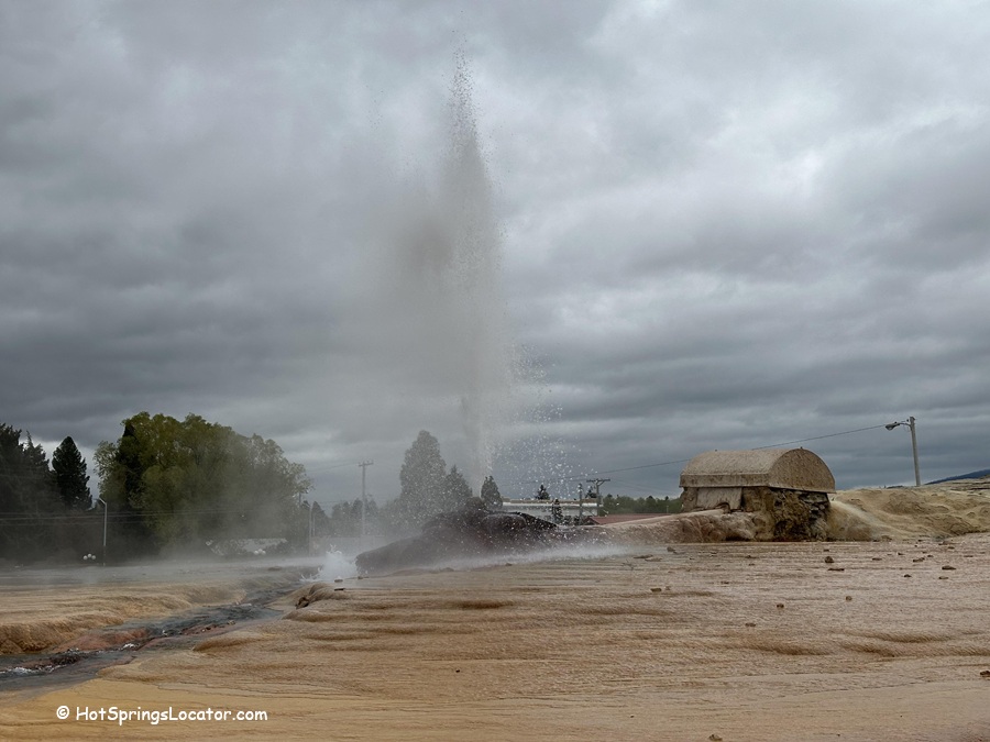 Soda Springs Geyser in Idaho - A close-up of the geyser eruption with water shooting high into the air against a cloudy sky. The foreground features mineral-rich ground, and a small structure is visible to the right in the background.