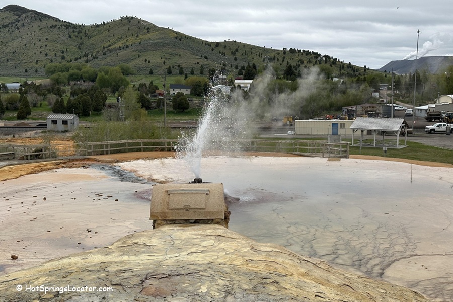 Soda Springs Geyser in Idaho - A view of the Soda Springs Geyser erupting, surrounded by a wooden fence and a small pool of mineral deposits. In the background, there are green hills, trees, and small town buildings under a cloudy sky.