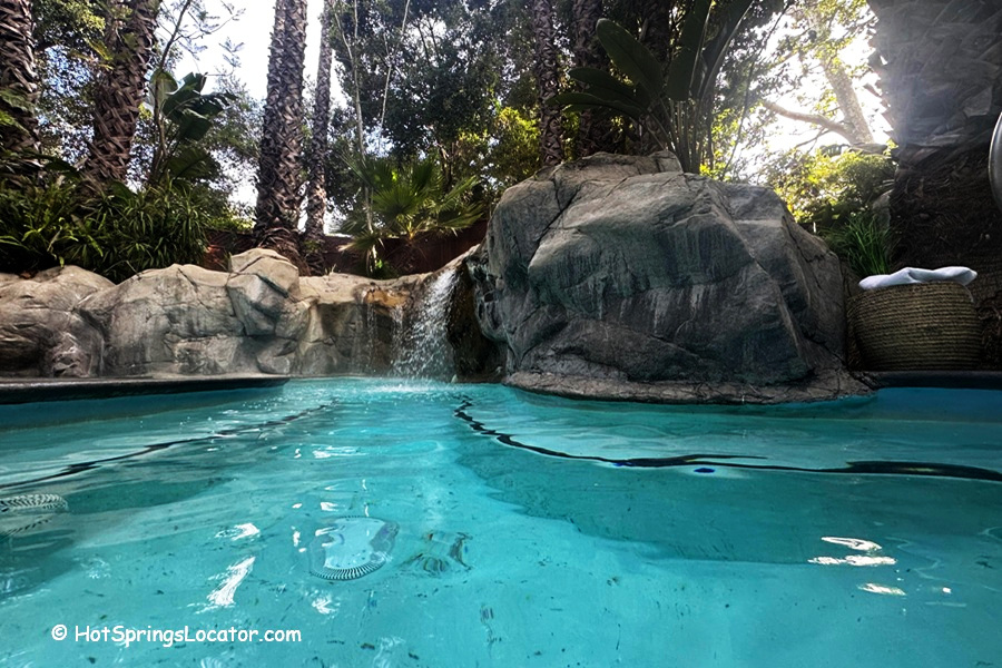 Sycamore Mineral Springs Resort & Spa California: The picture "Sycamore Mineral Springs Resort & Spa California hot tub with waterfall" shows a close-up view of a blue hot tub with a waterfall flowing from a large rock, surrounded by palm trees and lush vegetation.