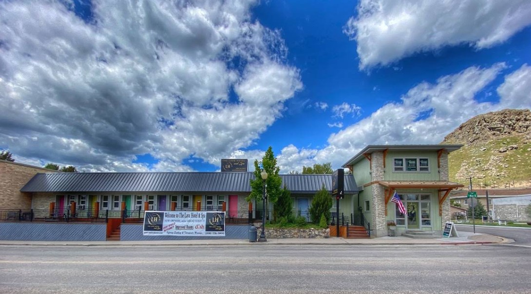The Lava Hotel & Spa idaho: The picture "The Lava Hotel & Spa idaho exterior" features a wide view of the hotel’s exterior, with colorful doors, a welcome banner, landscaping, and an American flag, under a dramatic blue sky with clouds and a hillside in the background.