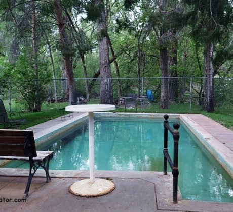 The Lava Hotel & Spa idaho: The picture "The Lava Hotel & Spa idaho hot springs pool with bench" shows a small rectangular hot springs pool surrounded by a patio area with a wooden bench and metal chairs, set in a shaded area with tall trees and a chain-link fence.