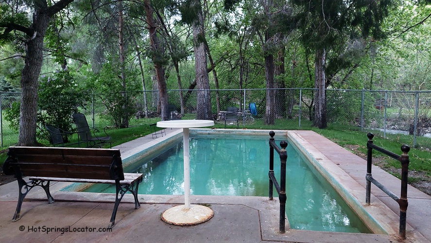 The Lava Hotel & Spa idaho: The picture "The Lava Hotel & Spa idaho hot springs pool with bench" shows a small rectangular hot springs pool surrounded by a patio area with a wooden bench and metal chairs, set in a shaded area with tall trees and a chain-link fence.