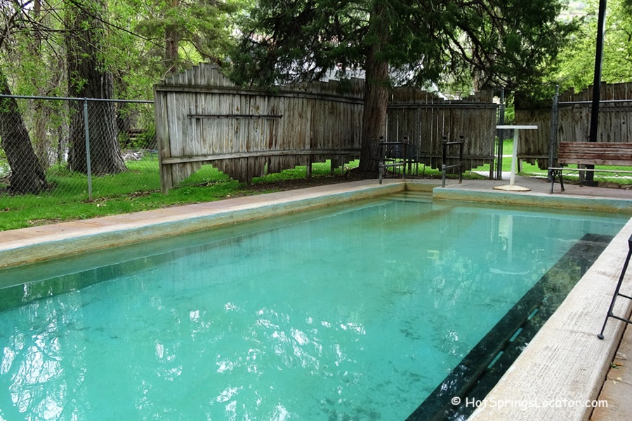 The Lava Hotel & Spa idaho: The picture "The Lava Hotel & Spa idaho hot springs pool" displays a clear, rectangular hot springs pool with turquoise water, bordered by a patio with seating, wooden fencing, and green grass, surrounded by tall trees.