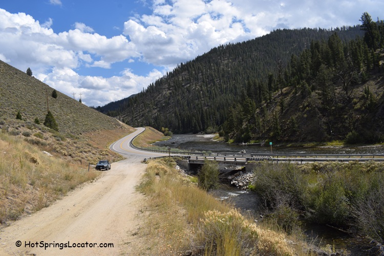 Basin Creek Hot Springs: The picture "Basin Creek Hot Springs sandy slope and creek" displays a sandy hillside with scattered rocks and dry plants sloping down to a creek, with dense green trees and forested mountains in the background.
