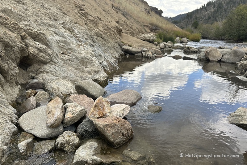 Basin Creek Hot Springs: The picture "Basin Creek Hot Springs rocky pool by the creek" shows a small hot spring pool lined with rocks at the edge of a sandy hillside, with clear water reflecting the sky and a flowing creek nearby in a mountain landscape.