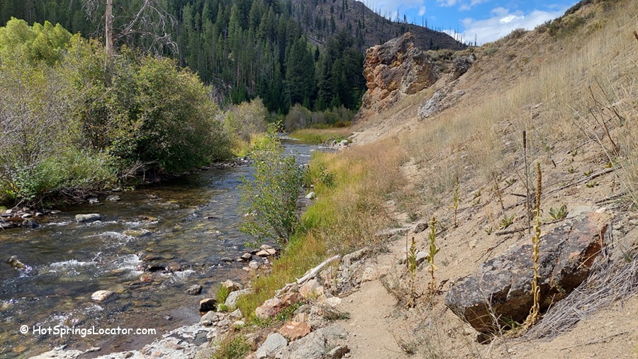 Basin Creek Hot Springs: The picture "Basin Creek Hot Springs road and creek bridge" shows a winding dirt road leading to a bridge over a creek, with a parked car, grassy hillsides, and a forested mountain under a partly cloudy sky.