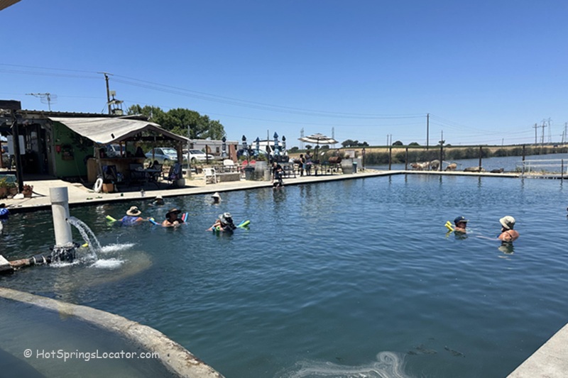 Franklin Hot Springs in California: The picture "Franklin Hot Springs in California pool with swimmers" displays an outdoor soaking pool with people floating and relaxing, a shaded green structure nearby, and open skies above the surrounding landscape.
