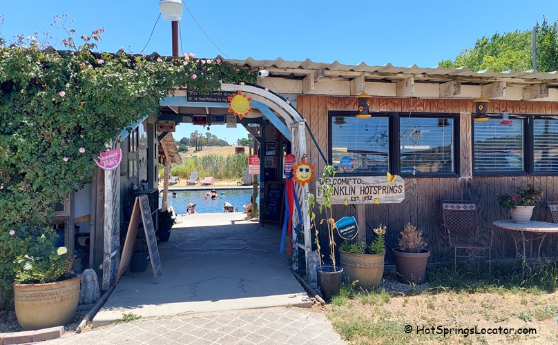 Franklin Hot Springs in California: The picture "Franklin Hot Springs in California entrance" shows a rustic wooden entrance with a sign welcoming visitors to Franklin Hot Springs, surrounded by potted plants, outdoor seating, and a view of the pool and reeds in the background.