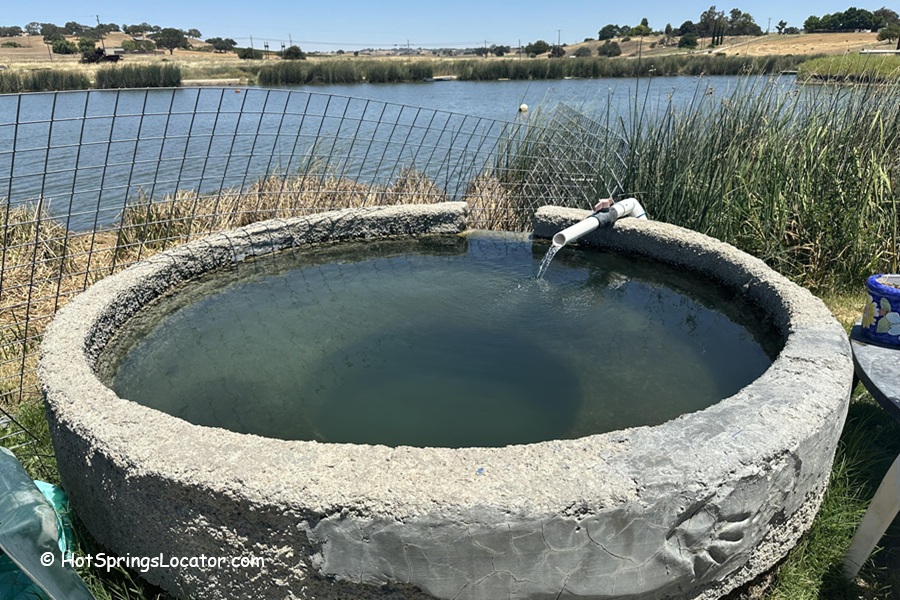 Franklin Hot Springs in California: The picture "Franklin Hot Springs in California round tub by lake" shows a small, circular concrete soaking tub filled with water, placed beside a wire fence and overlooking a lake and grassy landscape.