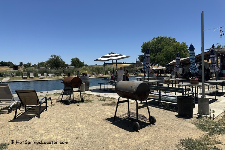 Franklin Hot Springs in California: The picture "Franklin Hot Springs in California picnic and barbecue area" features picnic tables with striped umbrellas and two barbecue grills set on a sandy area next to the soaking pool, with lounge chairs and grassy wetlands in the background.