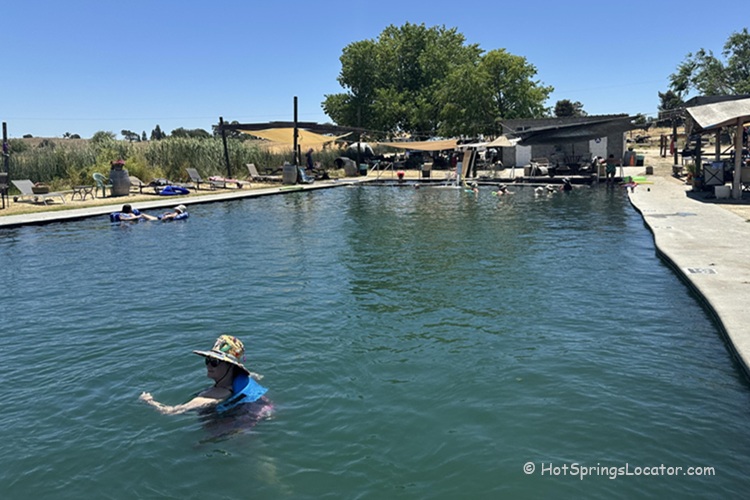 Franklin Hot Springs in California: The picture "Franklin Hot Springs in California main soaking pool" shows a large outdoor soaking pool with people swimming and relaxing, surrounded by a concrete deck, grassy area with lounge chairs, and shaded structures under a clear blue sky.