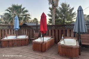 Mercey Hot Springs in California: The picture "Mercey Hot Springs in California private soaking tubs" displays three wooden soaking tubs with tall umbrellas on a wooden deck, surrounded by a privacy fence and palm trees in the background.
