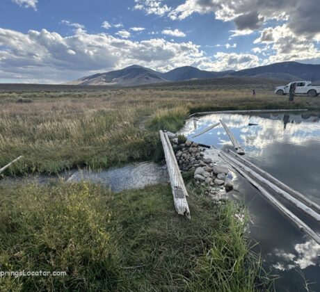 Barney Hot Springs in Idaho: The picture "Barney Hot Springs in Idaho natural pool with logs and trucks" shows a natural hot spring pool edged with wooden logs and rocks, with grassy meadows, several parked trucks, people in the distance, and mountains under a partly cloudy sky.