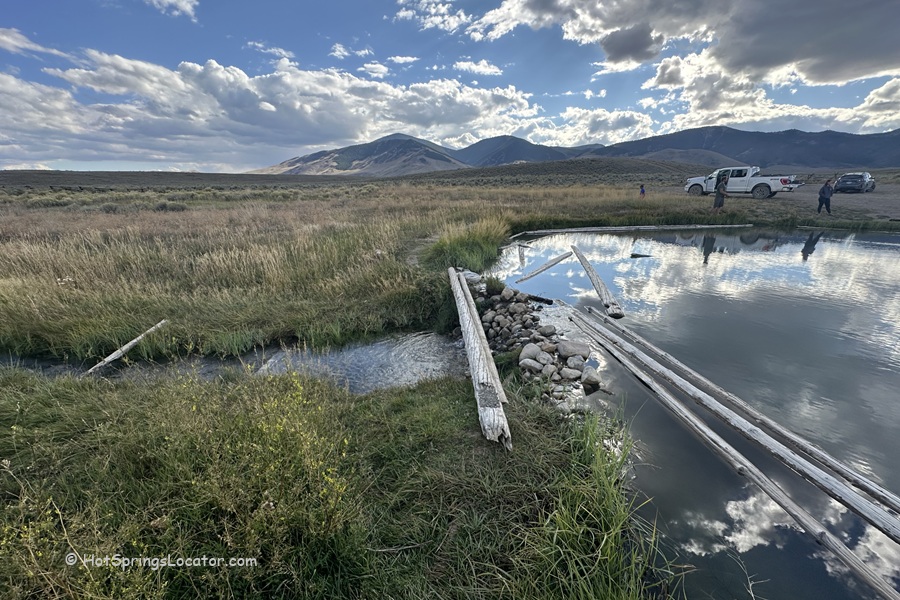 Barney Hot Springs in Idaho: The picture "Barney Hot Springs in Idaho natural pool with logs and trucks" shows a natural hot spring pool edged with wooden logs and rocks, with grassy meadows, several parked trucks, people in the distance, and mountains under a partly cloudy sky.