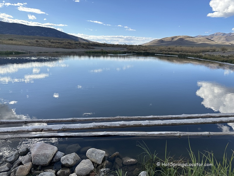 Barney Hot Springs in Idaho: The picture "Barney Hot Springs in Idaho hot spring pond with mountain view" displays a calm hot spring pond with logs and rocks along the shoreline, reflecting clouds in the water, surrounded by open grassland and rolling hills with mountains in the distance.
