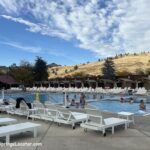 Kah-Nee-Ta Resort and Spa in Idaho: The picture "Kah-Nee-Ta Resort and Spa in Idaho outdoor swimming pool with lounge chairs and hills" shows people relaxing in a large outdoor swimming pool, surrounded by white lounge chairs and cabanas, with rolling golden hills and a partly cloudy sky in the background.