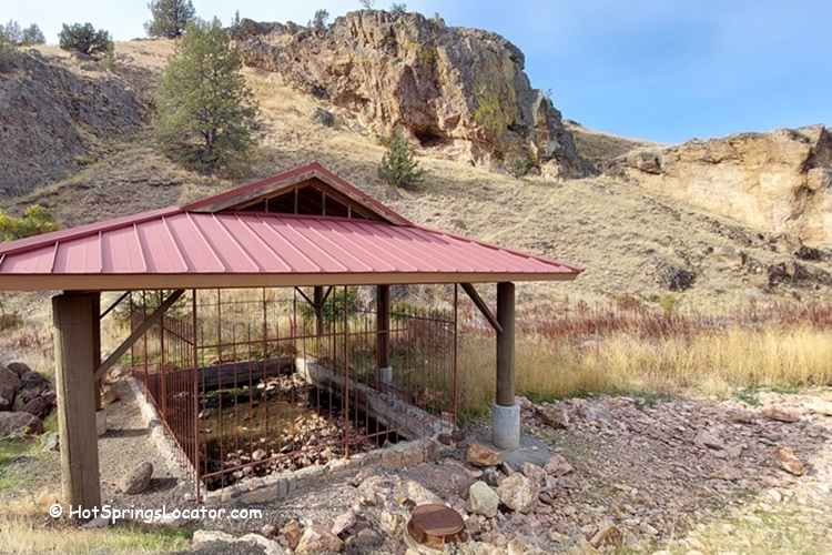 Kah-Nee-Ta Resort and Spa in Idaho: The picture "Kah-Nee-Ta Resort and Spa in Oregon hot spring source under red metal roof" displays a natural hot spring source protected by a red metal roof and surrounded by a fence, set against a backdrop of rocky hills and dry grass.