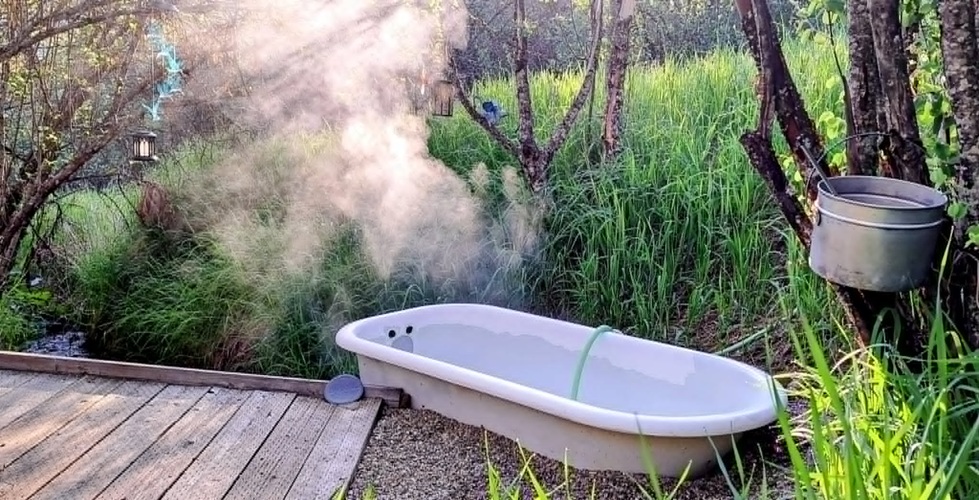 Double J&D Historic Hot Spring Ranch in Idaho: The picture "steaming outdoor bathtub in tall grass" shows a steaming bathtub placed on gravel next to a wooden deck, surrounded by tall green grass and trees, with sunlight streaming through the mist.
