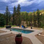 Double J&D Historic Hot Spring Ranch in Idaho: The picture "hot spring pool with lounge chair" captures a small rectangular hot spring pool on a gravel patio with a blue lounge chair and potted plant in the foreground, set against a backdrop of tall pine trees and dramatic cloudy sky.