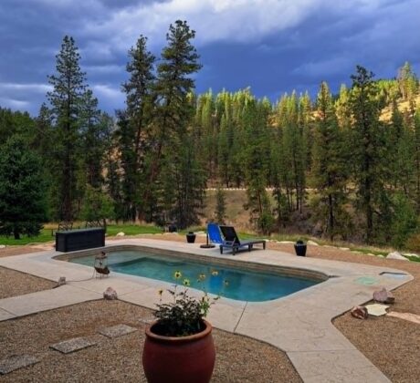 Double J&D Historic Hot Spring Ranch in Idaho: The picture "hot spring pool with lounge chair" captures a small rectangular hot spring pool on a gravel patio with a blue lounge chair and potted plant in the foreground, set against a backdrop of tall pine trees and dramatic cloudy sky.