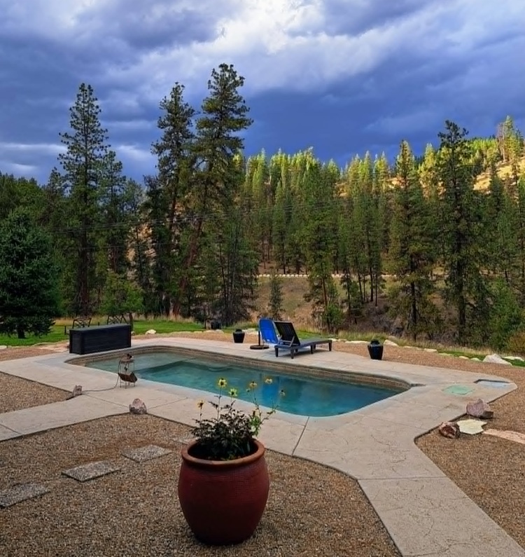 Double J&D Historic Hot Spring Ranch in Idaho: The picture "hot spring pool with lounge chair" captures a small rectangular hot spring pool on a gravel patio with a blue lounge chair and potted plant in the foreground, set against a backdrop of tall pine trees and dramatic cloudy sky.
