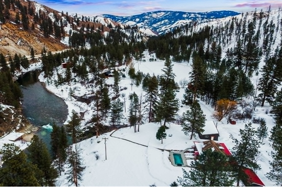 Double J&D Historic Hot Spring Ranch in Idaho: The picture "aerial view winter ranch setting" shows an aerial view of a hot spring ranch in a snowy mountain valley, with forested hills, a river, and outdoor pools surrounded by trees.